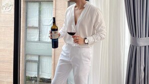 A person holding a wine bottle and glass, standing by a window with a view of a building. - Opera House Suites (Hanoi)