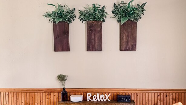 A wooden wall with three vertical planters containing green plants. Below, a shelf holds a vase with a small plant, a white container, and a black object. A wooden headboard with a woven seat is in front. - Des Moines River Front Cabin- Fishing/Hunting/Kayaking (Keosauqua)