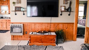 A living room with a large flat-screen TV mounted on the wall, wooden paneling on the walls, a wooden bench with a remote control, and a small air conditioning unit. - Des Moines River Front Cabin- Fishing/Hunting/Kayaking (Keosauqua)