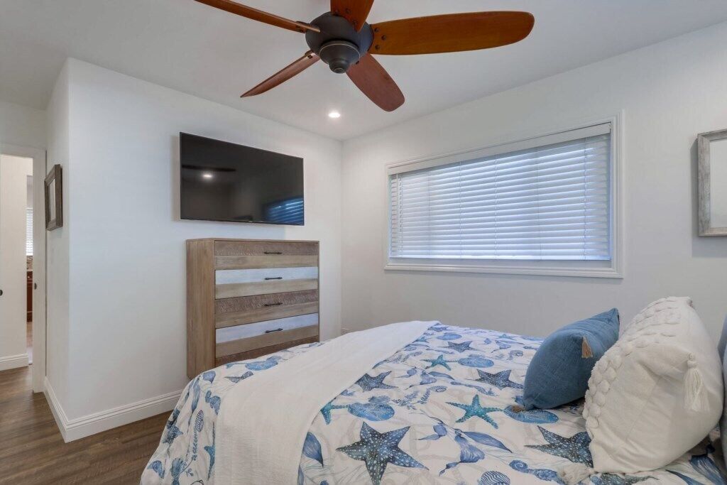 A bedroom with a bed, a ceiling fan, a dresser, a window with blinds, and a flat-screen TV mounted on the wall.