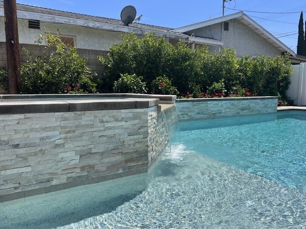 A swimming pool with stone walls, surrounded by greenery and a house in the background.