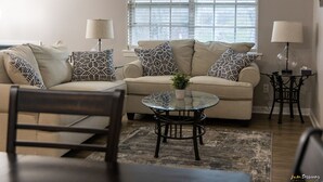 A living room with a beige sofa, patterned pillows, a glass coffee table, and two lamps.