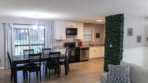 A modern kitchen with white cabinets, a microwave, and a coffee maker. There is a dining area with a table and chairs, and a living room with a gray sofa and patterned pillow.