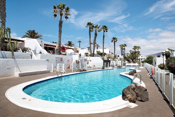 A swimming pool with a white pool deck, palm trees, and white buildings.