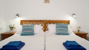 A double bed with white linens, two pillows, two blue towels, a wooden headboard, a small potted plant, and a wall-mounted light fixture.