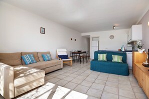 A living room with a beige sofa, a blue sofa, a wooden chair, a dining table, and a kitchen area with cabinets and a refrigerator. - Apartment 'Flamingo 62' with Private Terrace & Shared Pool (Playa Blanca)