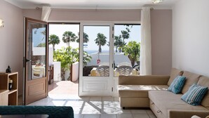 A living room with a sofa, a dining area with chairs and a table, and a sliding glass door leading to an outdoor patio with plants and a view of palm trees.