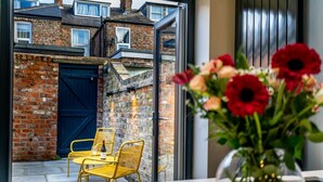 A view of a brick wall with a blue door, a yellow chair, and a table with a vase of flowers.