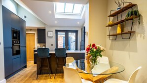 A modern kitchen with a skylight, a dining area with a glass table and chairs, and a built-in shelf with plants.