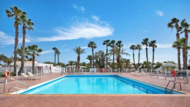 A rectangular swimming pool with clear blue water, surrounded by palm trees and a tiled deck. - Apartment 'Jardin del Sol 26' with Terrace, Shared Pool& Wi-Fi (Playa Blanca)