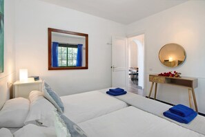 A hotel room with two beds, a bedside table with a lamp, a mirror, and a wooden console table with flowers.