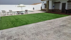 A pool area with white lounge chairs and a white umbrella, a well-maintained lawn, and a building with a tiled roof and stone base.