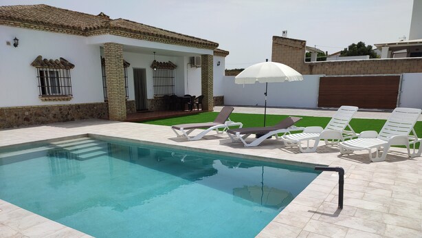 A swimming pool with lounge chairs and an umbrella, surrounded by a white building with a tiled roof.