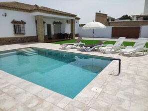 A swimming pool with lounge chairs and an umbrella, surrounded by a white building with a tiled roof.