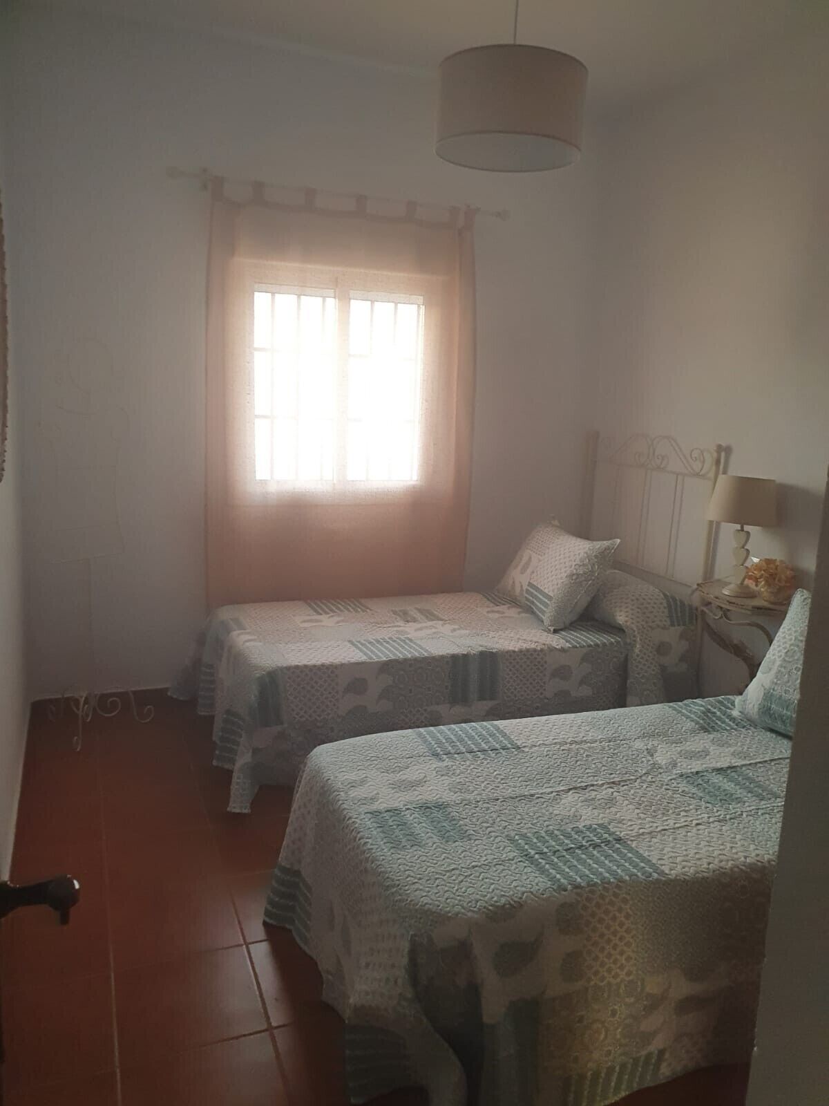 A room with two beds, a window with curtains, a hanging lamp, and a small table with a lamp.