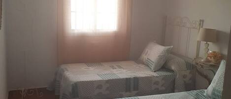 A room with two beds, a window with curtains, a hanging lamp, and a small table with a lamp.