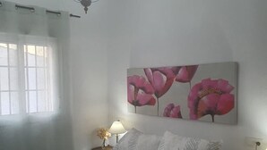 A bedroom with a bed, two bedside tables, a hanging light fixture, a window with curtains, and a wall-mounted painting of flowers.