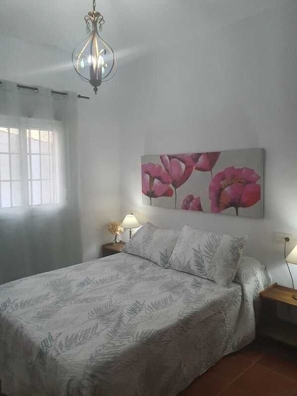 A bedroom with a bed, two bedside tables, a hanging light fixture, a window with curtains, and a wall-mounted painting of flowers.