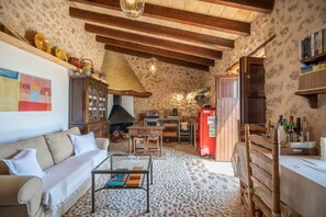 Un interior rústic amb una paret de pedra, bigues de fusta, un sofà, una taula de cafè, una llar de foc, una zona de cuina i una prestatgeria.