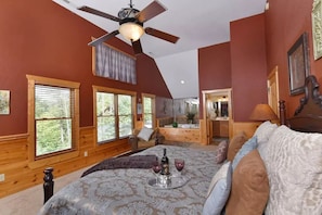 A bedroom with a large bed, a ceiling fan, wooden walls, and a view of the outdoors through the windows.