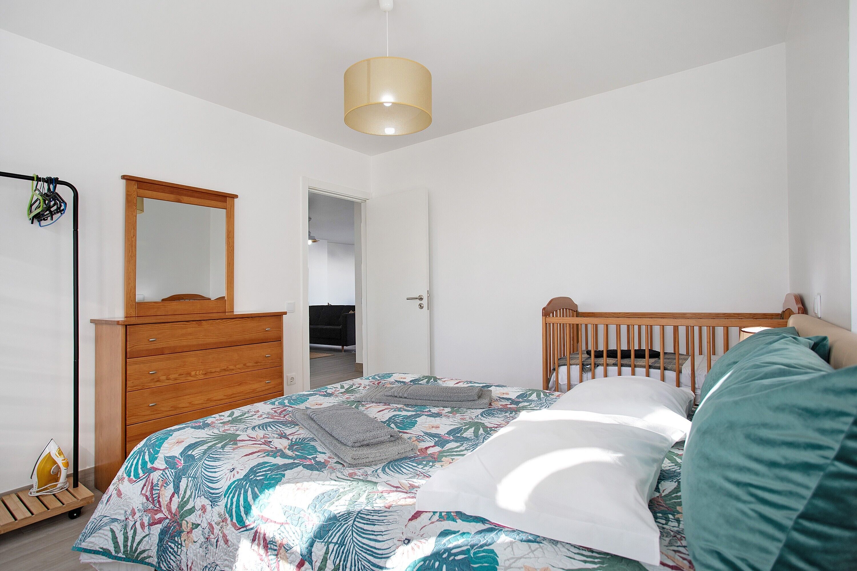 Ein Schlafzimmer mit einem Bett, einer Holzkommode, einem Spiegel, einem Babybett und einer Deckenleuchte.