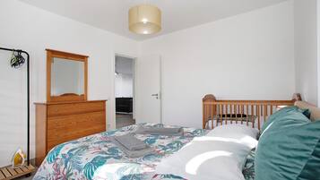 Ein Schlafzimmer mit einem Bett, einer Holzkommode, einem Spiegel, einem Babybett und einer Deckenleuchte.