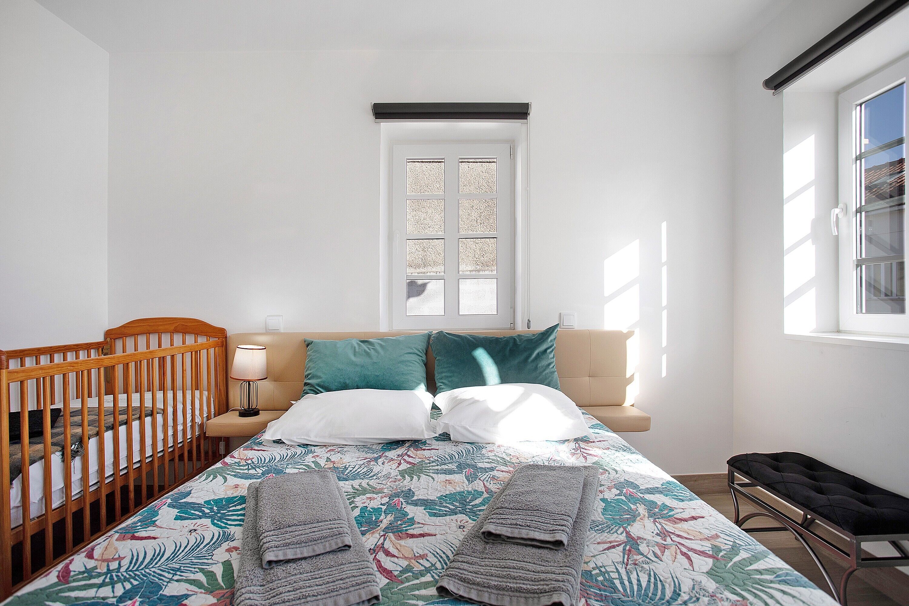 Une chambre avec un berceau, un lit avec une literie à motifs floraux, un banc et une fenêtre avec des stores.