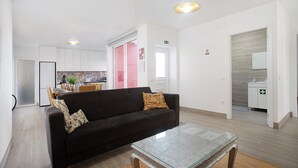 Una sala de estar moderna con un sofá negro, una mesa de centro de vidrio y una cocina al fondo.