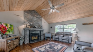 Un salon chaleureux avec une cheminée, un ventilateur de plafond, une grande fenêtre et un coin salon confortable.