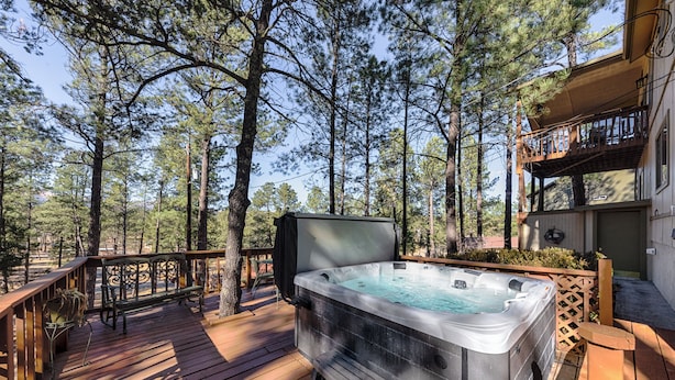 Un bain à remous sur une terrasse en bois, entouré de grands pins, avec une balustrade en bois et un bâtiment doté d’un balcon en arrière-plan.