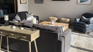 A modern living room with a grey sofa, a wooden coffee table, and a ceiling fan. There are framed pictures on the wall and a flat-screen TV.