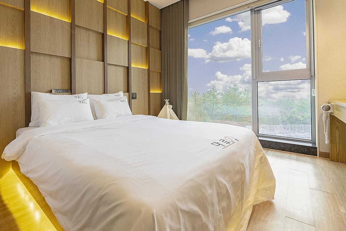 Une chambre moderne avec un grand lit, des murs recouverts de panneaux en bois et une vue sur les arbres par une grande fenêtre.