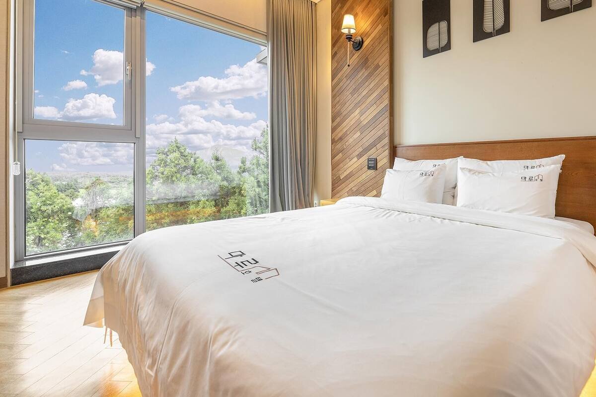 Une chambre d’hôtel dotée d’une grande fenêtre donnant sur des arbres et des montagnes, d’une tête de lit en bois et d’un lit avec une literie blanche.