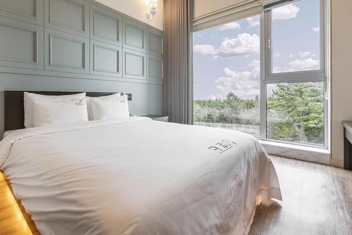 Une chambre avec une grande fenêtre, un lit avec une literie blanche, et une vue sur les arbres et le ciel.