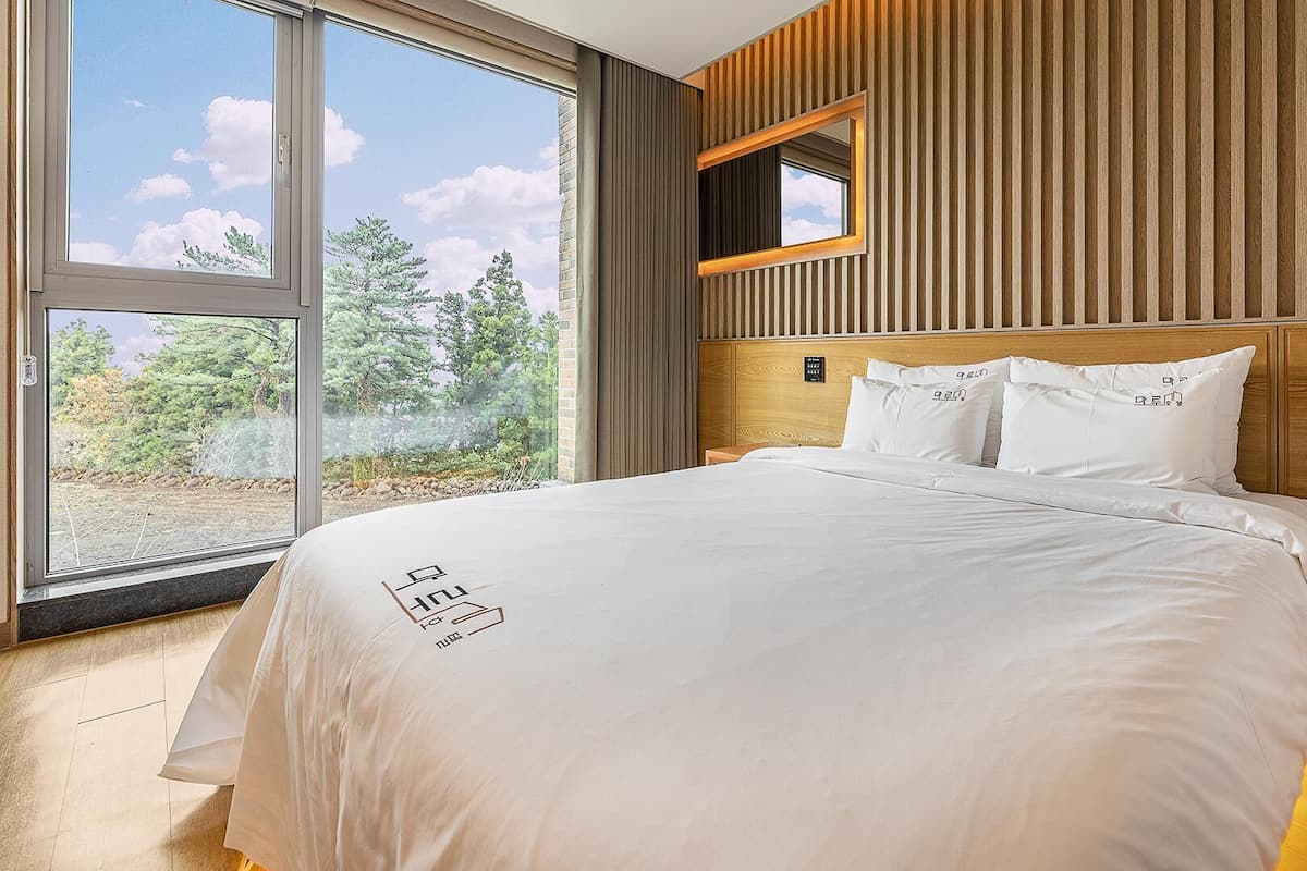 Une chambre moderne avec une grande fenêtre, une tête de lit en bois et un lit recouvert de linge de maison blanc.
