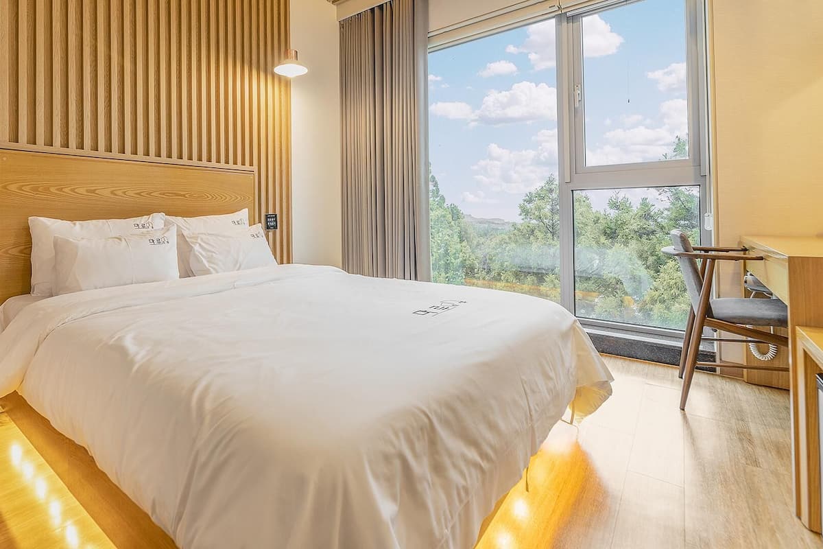 Une chambre d’hôtel moderne dotée d’un grand lit, de murs recouverts de panneaux de bois, d’un bureau avec une chaise et d’une grande fenêtre offrant une vue panoramique.