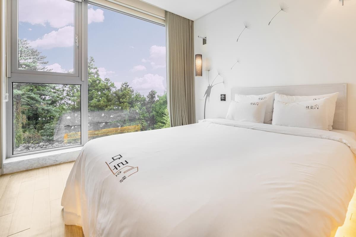 Une chambre moderne avec une grande fenêtre, un lit recouvert de linge de lit blanc et une vue sur les arbres et le ciel.