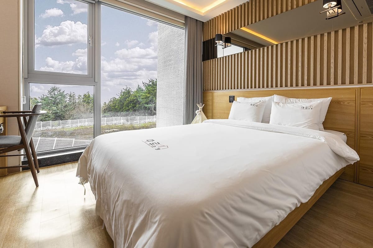 Une chambre d’hôtel moderne avec un grand lit, une tête de lit en bois et une vue sur l’extérieur grâce à de grandes fenêtres.