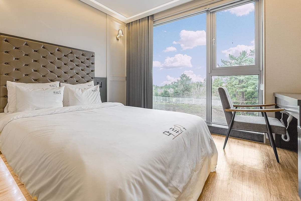 Une chambre d’hôtel moderne dotée d’un grand lit, d’une tête de lit au motif capitonné, d’un bureau avec une chaise et offrant une vue sur l’extérieur grâce à une grande fenêtre.