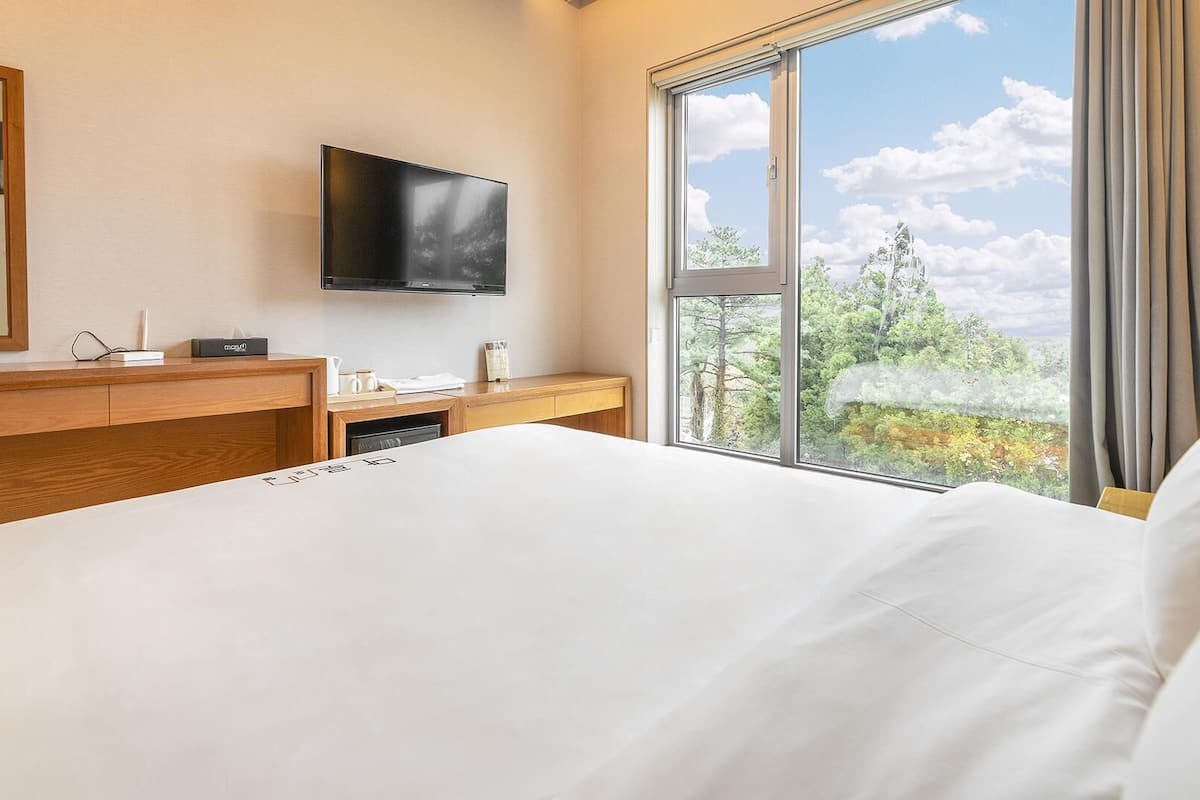 Une chambre d’hôtel équipée d’un lit, d’un bureau, d’un téléviseur à écran plat fixé au mur et d’une grande fenêtre donnant sur un paysage de verdure et de ciel.