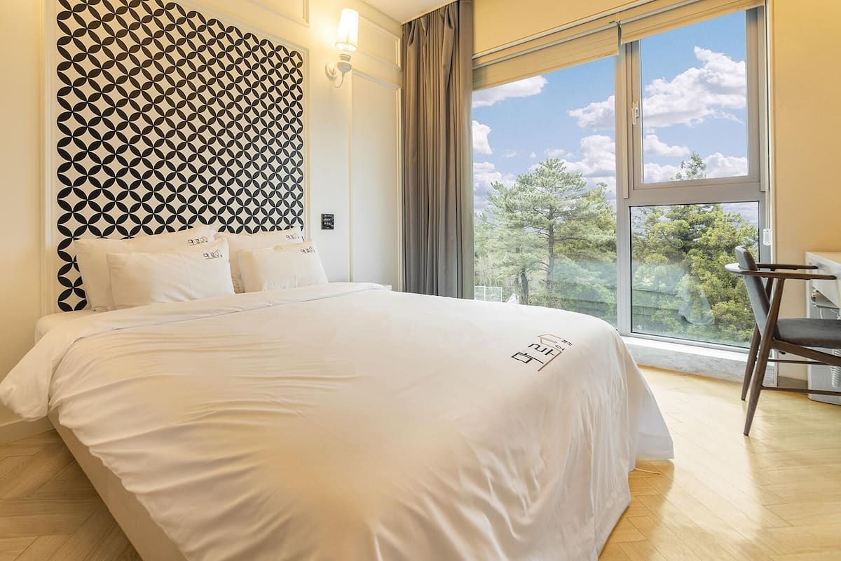 Une chambre d’hôtel moderne dotée d’un grand lit, d’un bureau avec une chaise et offrant une vue sur les arbres à travers une grande fenêtre.
