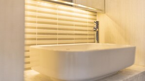 Ein Badezimmer mit einem weißen Waschbecken, einem chromfarbenen Wasserhahn und einem Fenster mit horizontalen Rollläden.