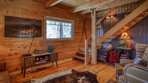 Interior acogedor de una cabaña con suelo de madera, un televisor grande, una ventana, una escalera y una zona de estar con un manto de piel.