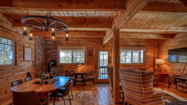 Interior rústico de una cabaña con una mesa de comedor de madera, sillas, un sillón a rayas, una chimenea y un televisor.