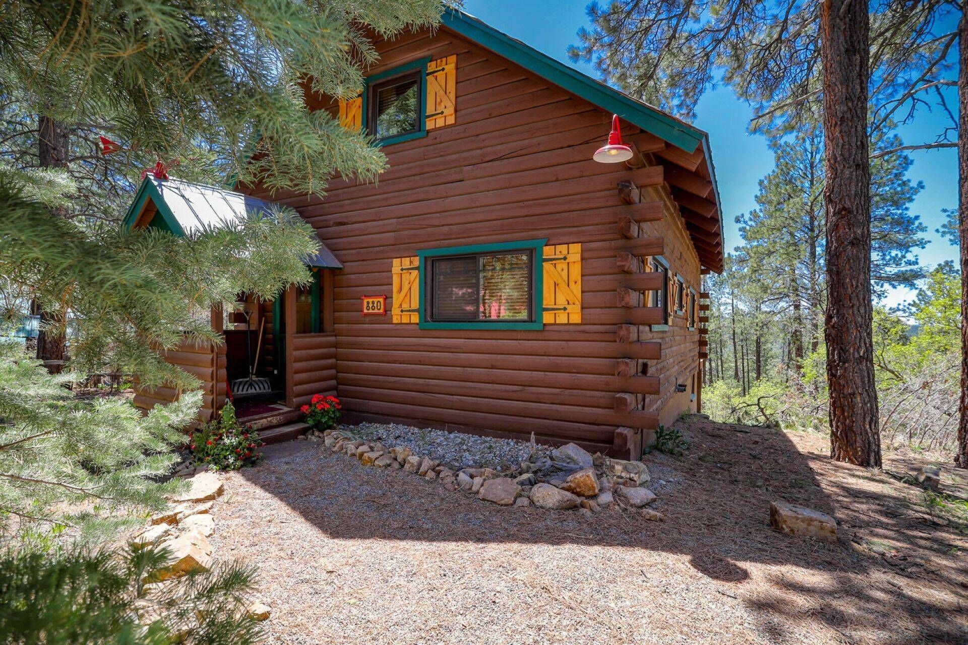 Une cabane rustique en rondins de bois, avec un toit végétalisé, un petit porche et une allée de gravier entourée d’arbres.