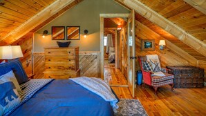 Un dormitorio acogedor en el ático con techo de madera, cama con ropa de cama azul, cómoda, una silla y una mesita.