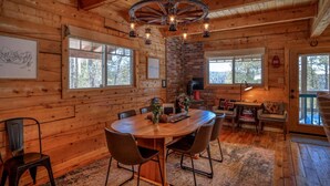 Interior de una cabaña de madera con una mesa redonda para comer, sillas, chimenea y un ventanal grande.