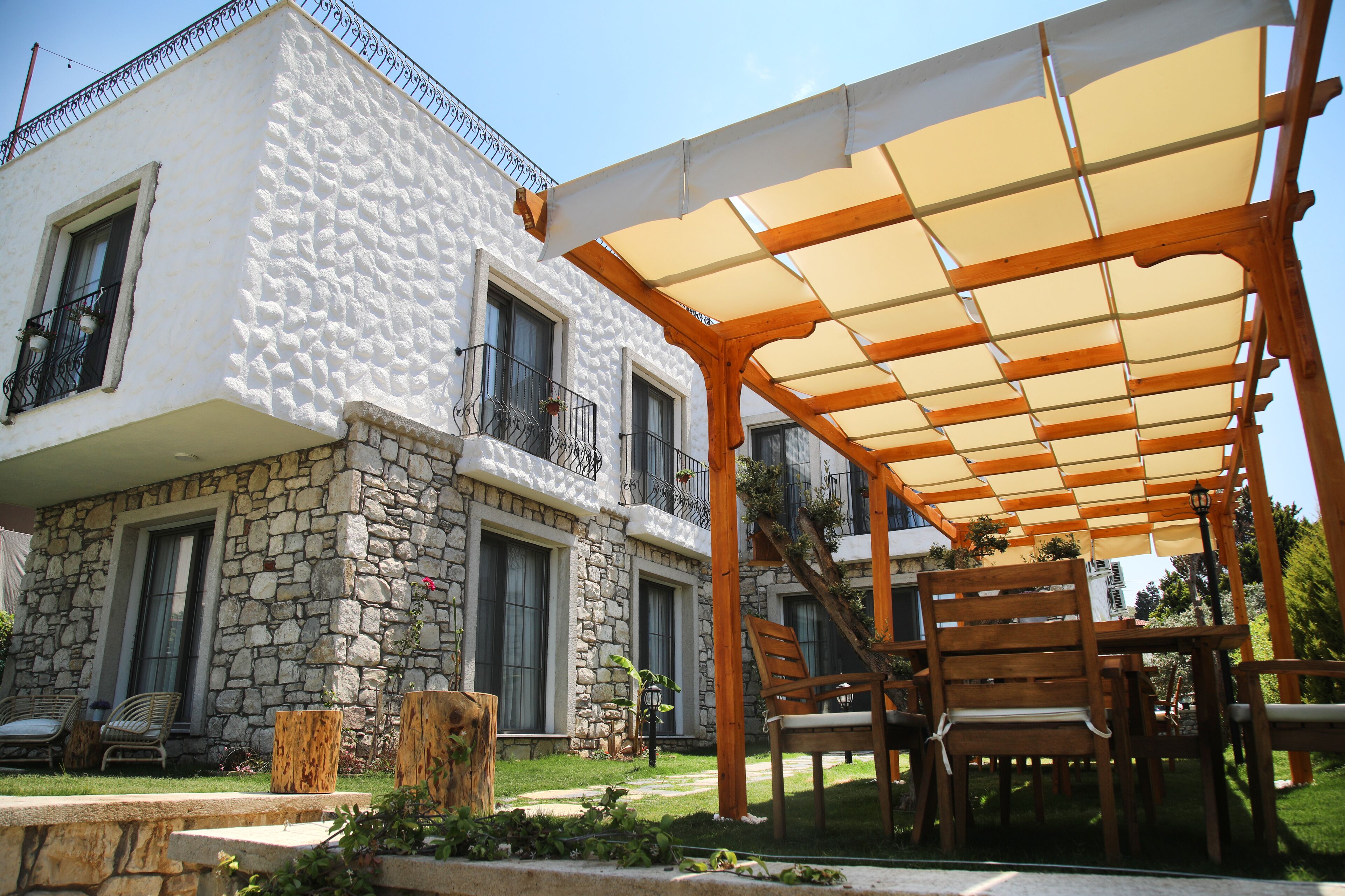 Un edificio de piedra con balcón, una pérgola de madera y mobiliario de exterior.