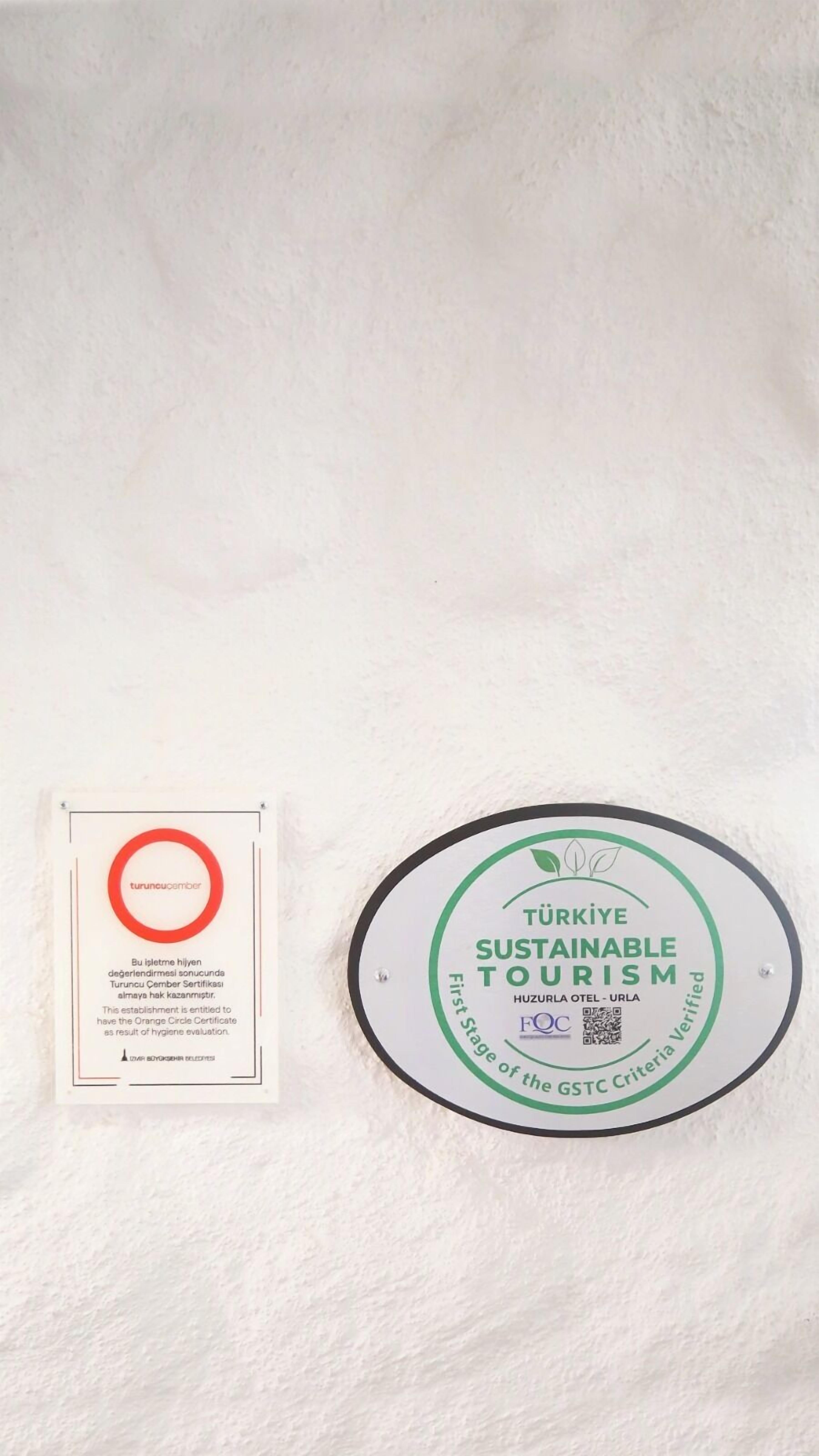Eine weiße Wand mit zwei Schildern. Ein Schild ist ein roter Kreis mit dem Text „turuncucember“ und einer Beschreibung in einer anderen Sprache. Das andere Schild ist ein grüner Kreis mit dem Text „TÜRKIYE SUSTAINABLE TOURISM“ und einem QR-Code.