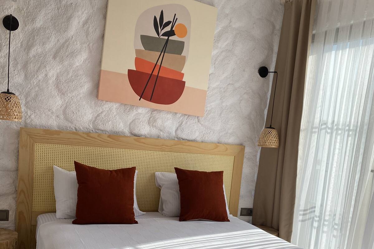 A hotel room with a large bed, a headboard, a wall-mounted artwork, and a window with curtains.